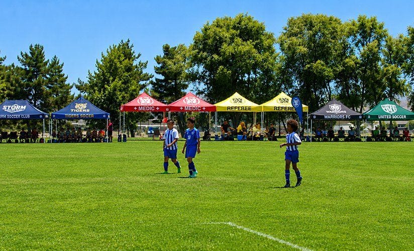Branded canopy tents providing sideline shade for youth sports teams during a soccer tournament, showcasing team branding and outdoor event shelter solutions.